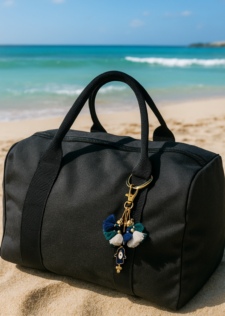 Black duffel bag with a decorative keychain on a sandy beach with ocean in the background